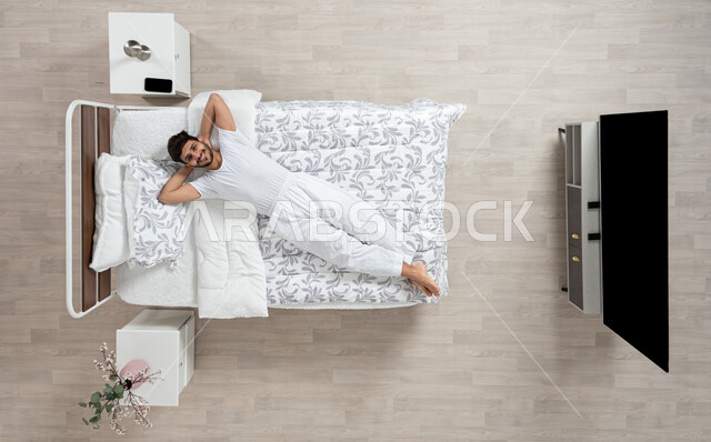 الاسترخاء والراحة وقت القيلوله، بورتريه علوي عامودي لشاب عربي خليجي سعودي يرتدي الفانيلا والسروال الابيض مستلقي على سريره واضعا يديه خلف رأسه، ايماءات وجه ويدين تدل على السعادة، الاستمتاع بنوم هادئ وصحي، وسادة بيضاء ناعمة، خلفيه بيج