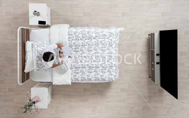 القراءة و الإطلاع و الاستعداد للعالم الدراسي الجديد، صورة علوية عاموديه لشاب عربي خليجي سعودي يقوم بقراءة كتاب جالسا على سريره مرتديا الفانيلا والسروال الابيض الداخلي، كتب علمية وروايات ثقافية، حب المطالعة و التثقيف، تنمية المهارات الذهنية و ممارسة هواية القراءه، خلفيه بيج