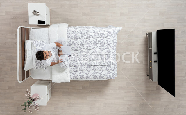 Reading, learning, and preparing for the new academic world, a vertical overhead picture of a Saudi Gulf Arab young man reading a book sitting on his bed, wearing a flannel and white underpants, looking up with a smile, scientific books and cultural novels, developing mental skills and practicing the hobby of reading, beige background.
