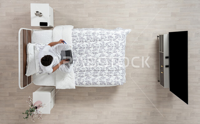 Performing duties and working remotely, using modern devices for browsing and chatting, a vertical overhead portrait of a Saudi Gulf Arab young man wearing flannel and white underpants, working on his laptop, sitting on his bed and holding his mobile phone in his other hand, beige background
