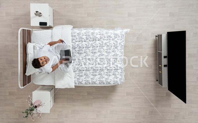 أداء الواجبات والعمل عن بعد ، استخدام الاجهزه الحديثه في التصفح والدردشه ، بورتريه علوي عامودي لشاب عربي خليجي سعودي يرتدي الفانيلا والسروال الداخلي الابيض يعمل على جهاز الكمبيوتر المحمول جالسا على سريره ويمسك بيده الاخرى هاتفه المحمول ينظر الى الاعلى بابتسامه، خلفية بيج