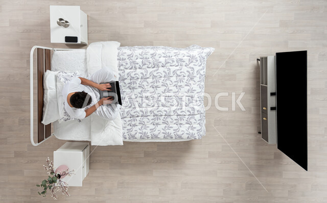 Performing duties and working remotely, using the laptop for browsing and chatting, a vertical overhead portrait of a young Saudi Gulf Arab man wearing flannel and white underpants, working on the laptop sitting on his bed, beige background