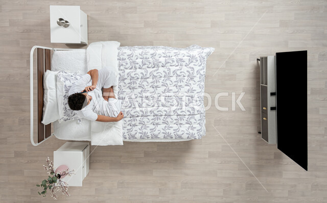 إجراء مكالمات هاتفية باستخدام الهاتف المحمول،  دردشة ومحادثات والتواصل مع الأفراد ، بورتريه علوي عامودي لشاب عربي خليجي سعودي يرتدي الفانيلا والسروال الداخلي الابيض  يقوم باجراء مكالمه بجهازه المحمول جالسا على سريره، خلفية بيج