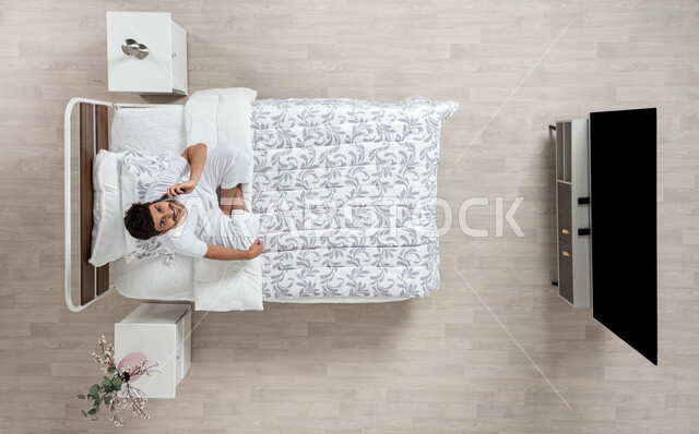 إجراء مكالمات هاتفية باستخدام الهاتف المحمول،  دردشة ومحادثات والتواصل مع الأفراد ، بورتريه علوي عامودي لشاب عربي خليجي سعودي يرتدي الفانيلا والسروال الداخلي الابيض مبتسم ينظر الى الاعلى يقوم باجراء مكالمه بجهازه المحمول جالسا على سريره، خلفية بيج