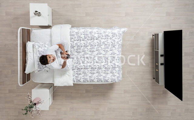 إجراء مكالمات هاتفية باستخدام الهاتف المحمول ,  دردشة ومحادثات والتواصل مع الأفراد ، بورتريه علوي عامودي لشاب عربي خليجي سعودي مبتسم ينظر الى الاعلى  يرتدي الفانيلا والسروال الداخلي الابيض يمسك بجهازه المحمول جالسا على سريره، خلفية بيج