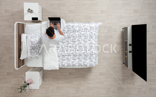 أداء الواجبات والعمل عن بعد ، استخدام جهاز الاب التوب في التصفح والدردشه ، بورتريه علوي عامودي لشاب عربي خليجي سعودي يرتدي الفانيلا والسروال الداخلي الابيض يعمل على جهاز الكمبيوتر المحمول جالسا على سريره، خلفية بيج