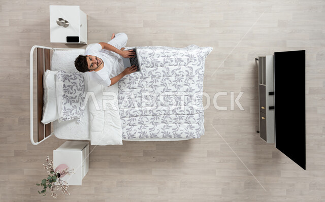 أداء الواجبات والعمل عن بعد ، استخدام جهاز الاب التوب في التصفح والدردشه ، بورتريه علوي عامودي لشاب عربي خليجي سعودي مبتسم ينظر الى الاعلى  يرتدي الفانيلا والسروال الداخلي الابيض يعمل على جهاز الكمبيوتر المحمول جالسا على سريره، خلفية بيج