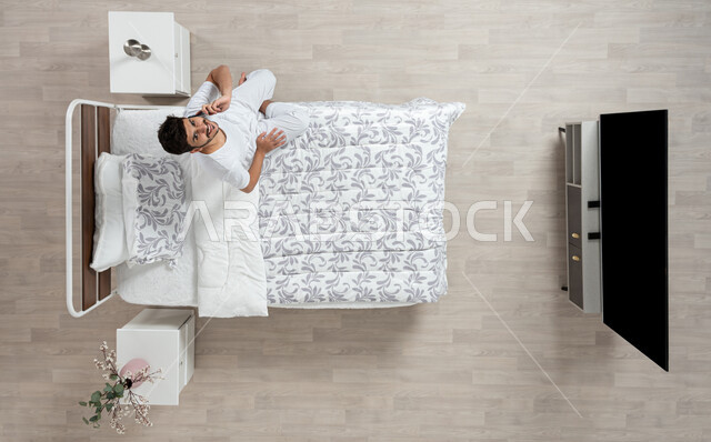 إجراء مكالمات هاتفية باستخدام الهاتف المحمول ,  دردشة ومحادثات والتواصل مع الأفراد ، بورتريه علوي عامودي لشاب عربي خليجي سعودي مبتسم ينظر الى الاعلى  يرتدي الفانيلا والسروال الداخلي الابيض يتحدث بجهازه المحمول على سريره، خلفية بيج