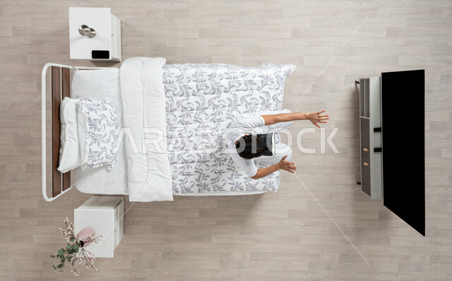 vertical portrait of a Saudi Gulf Arab young man wearing flannel and white underpants, sitting on his bed and watching TV with virtual reality (VR) technology using special glasses, pointing with his hands at the TV, using the sense of touch to move the holographic elements, beige background