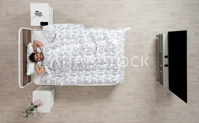 الاسترخاء والراحة وقت القيلوله، بورتريه علوي عامودي لشاب عربي خليجي سعودي يستلقي على السرير ويضع يده خلف راسه، ايماءات وجه ويدين تدل على السعادة، الاستمتاع بنوم هادئ وصحي، وسادة بيضاء ناعمة، النوم المبكر، خلفيه بيج