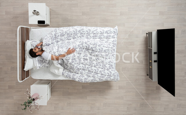 التعب والحاجة إلى الراحة بعد العمل, الشعور بالنعاس الشديد والرغبه في النوم ، بورتريه علوي عامودي لشاب عربي خليجي سعودي مستلقي في سريره يقوم بالتثاؤب بإيماءات التعب والارهاق، مفهوم الاسترخاء على سرير ناعم، خلفيه بيج