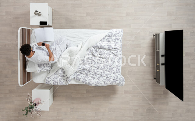 Reading and reading before bed, doing work in bed, an overhead portrait of a Saudi Gulf Arab man lying on his bed holding a cup of coffee looking at an open book, browsing books in the bedroom, doing tasks in bed, consuming stimulants and hot drinks, beige background