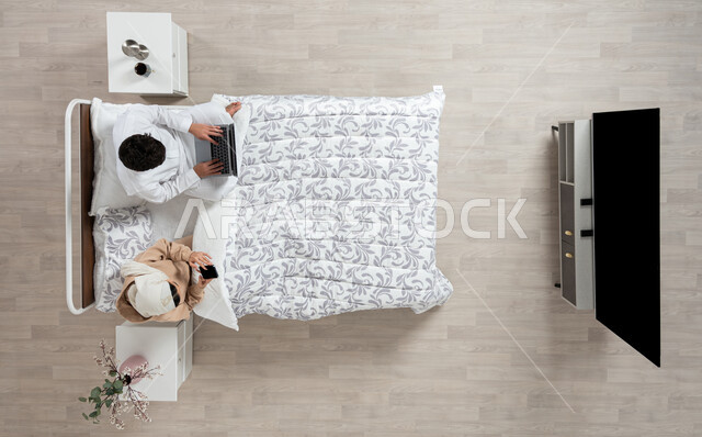 مفهوم التقنيات الحديثه والتواصل عن بعد، بورتريه عمودي علوي لسيده عربيه خليجيه سعوديه تجلس بجانب زوجها على سريرهما تمسك بيدها جهاز الهاتف المحمول ويقوم زوجها باستخدام الكمبيوتر الشخصي المحمول، التواصل مع الاهل والاصدقاء، العمل عن بعد في غرفه النوم، خلفيه بيج