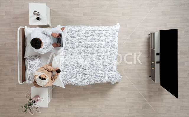 مفهوم التقنيات الحديثه والتواصل عن بعد، بورتريه عمودي علوي لسيده عربيه خليجيه سعوديه تجلس بجانب زوجها على سريرهما تمسك بيدها جهاز الهاتف المحمول ويقوم الزوج باستخدام الكمبيوتر الشخصي المحمول يقومان بالتحدث، العمل و اداء الواجبات في غرفه النوم، خلفيه بيج