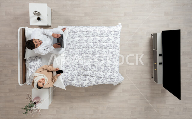 The concept of modern technologies and remote communication, an overhead vertical portrait of a Saudi Arabian Gulf woman sitting next to her husband on their bed, holding a mobile phone in her hand, and the husband using the laptop, looking up with a smile, working and performing duties in the bedroom, beige background.