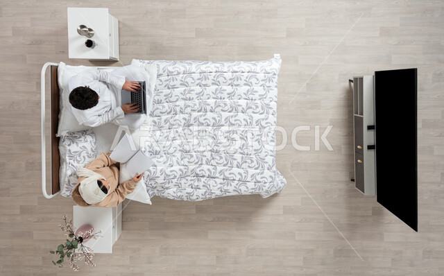 The concept of modern technologies and remote communication, an overhead vertical portrait of a Saudi Gulf Arab woman sitting next to her husband on their bed, holding an open book in her hand and the husband using the laptop, rest and relaxation in the bedroom, beige background