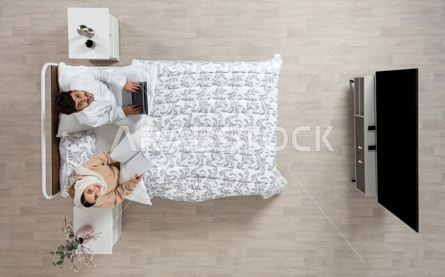 مفهوم التقنيات الحديثه والتواصل عن بعد، بورتريه عمودي علوي لسيده عربيه خليجيه سعوديه تجلس بجانب زوجها على سريرهما تمسك بيدها كتابا مفتوح ويقوم الزوج باستخدام الكمبيوتر الشخصي المحمول ينظران الى الاعلى بابتسامه، خلفيه بيج