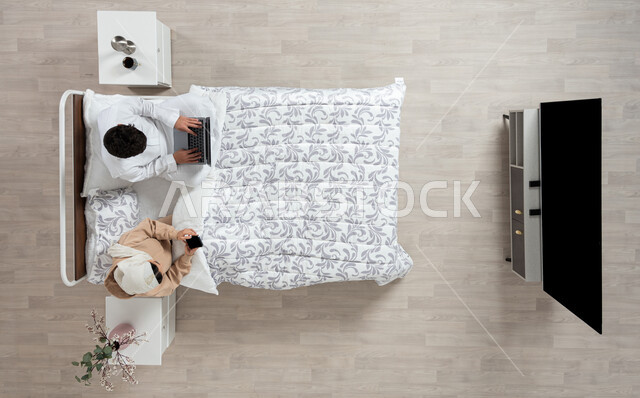 مفهوم التقنيات الحديثه والتواصل عن بعد، بورتريه عمودي علوي لسيده عربيه خليجيه سعوديه تجلس بجانب زوجها على سريرهما تمسك بيدها جهاز الهاتف المحمول ويقوم زوجها باستخدام الكمبيوتر الشخصي المحمول، التواصل مع الاهل والاصدقاء، خلفيه بيج