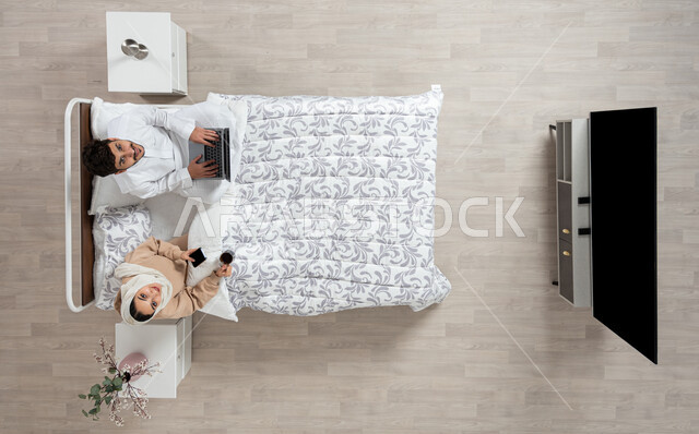 مفهوم التقنيات الحديثه والتواصل عن بعد، بورتريه عمودي علوي لسيده عربيه خليجيه سعوديه تجلس بجانب زوجها على سريرهما تمسك بيدها جهازها المحمول وكوب القهوه ويقوم زوجها باستخدام الكمبيوتر الشخصي المحمول ينظران الى الاعلى بابتسامه، التواصل مع الاهل والاصدقاء، خلفيه بيج