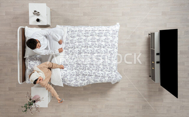 التقاط الصور والذكريات، بورتريه عمودي علوي لسيده عربيه خليجيه سعوديه تجلس بجانب زوجها على سريرهما تمسك بيدها جهازها المحمول وتقوم بالتقاط سيلفي، التواصل مع الاهل والاصدقاء، مفهوم التقنيات الحديثه، قضاء اوقات ممتعه، خلفيه بيج