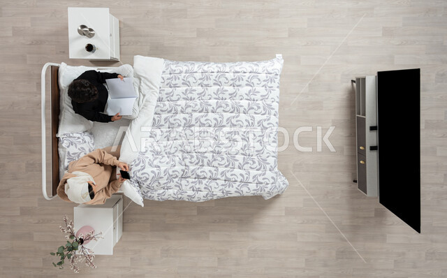 الاهتمام بتربيه ورعايه الابناء، بورتريه عمودي علوي لسيده عربيه خليجيه سعوديه تجلس على سريرها تمسك بيدها جهازها المحمول وبجانبها ابنها البالغ يدرس، التواصل مع الاهل والاصدقاء، اهميه الدراسه والتعلم، المذاكره والحفظ، خلفيه بيج