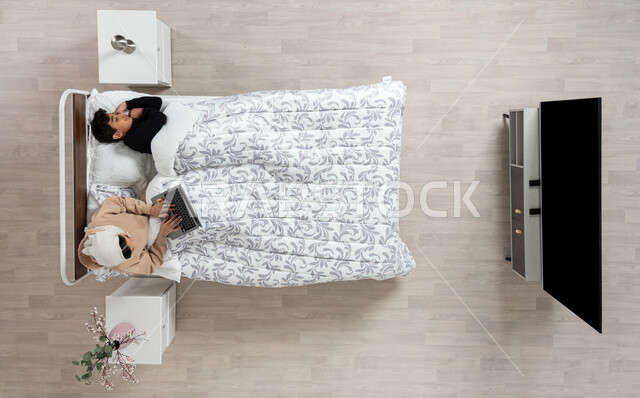 النوم بعمق من الارهاق والتعب، بورتريه عمودي علوي لسيده عربيه خليجيه سعوديه محجبه تجلس على سريرها بجوار ابنها النائم وتقوم باستخدام جهاز الاب توب المحمول، اخذ قيلولة و قسط من الراحة، الكسل والخمول والحاجة الى النوم، خلفيه بيج