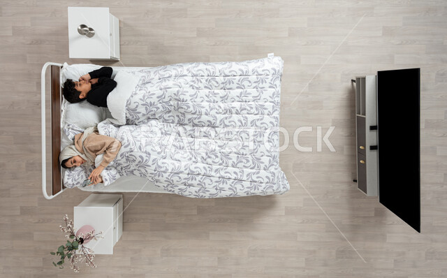 النوم بعمق من الارهاق والتعب، بورتريه عمودي علوي لسيده عربيه خليجيه سعوديه محجبه مستلقيه على سريرها بجوار ابنها، تمسك بيدها جهازها المحمول، اخذ قيلولة و قسط من الراحة، الكسل والخمول والحاجة الى النوم، خلفيه بيج