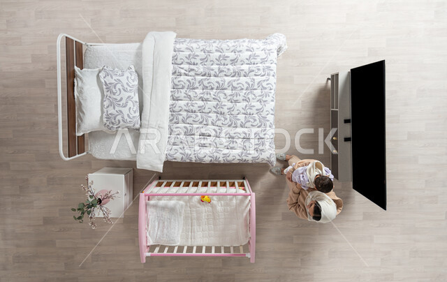 حنان الأمهات و تحمل المسؤولية والعناية بالأطفال، قضاء الوقت باللعب مع الأبناء بإيماءات السعادة والسرور،  بورتريه عامودي علوي لأم عربية خليجية سعودية محجبه تحمل طفلتها الرضيعة وتقوم بمداعبتها بحب وحنان في غرفه نومهما, خلفية بيج