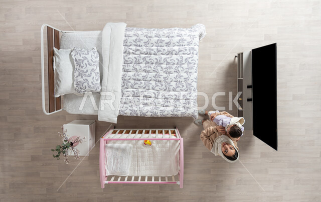 Mothers’ tenderness, taking responsibility and taking care of children, spending time playing with children with gestures of happiness and pleasure, overhead vertical portrait of a veiled Saudi Arabian Gulf mother carrying her infant daughter and caressing her with love and tenderness in their bedroom, looking up with a smile, beige background
