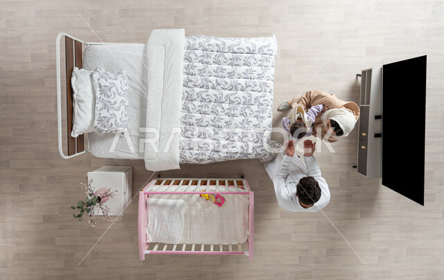 مداعبه الأطفال بحب وحنان، قضاء الوقت باللعب مع الأبناء بإيماءات السعادة والسرور، بورتريه علوي عامودي لأم واب عربيان خليجيان سعوديان يداعبان  طفلتهم الرضيعة وتقوم الام بحملها في غرفه نومهما، مفهوم الرعايه بالطفل, خلفية بيج