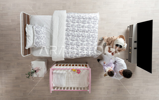 مداعبه الأطفال بحب وحنان، قضاء الوقت باللعب مع الأبناء بإيماءات السعادة والسرور، بورتريه علوي عامودي لأم واب عربيان خليجيان سعوديان يداعبان  طفلتهم الرضيعة ويقوم الاب بحملها في غرفه نومهما، مفهوم الرعايه بالطفل, خلفية بيج