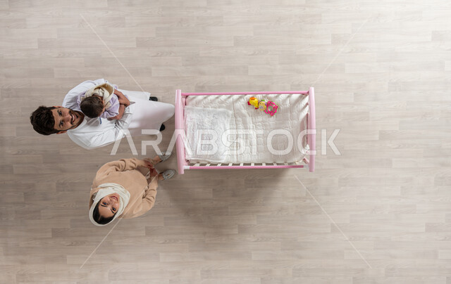 مداعبه الأطفال بحب وحنان، قضاء الوقت باللعب مع الأبناء بإيماءات السعادة والسرور، بورتريه علوي عامودي لأم واب عربيان خليجيان سعوديان ينظران بابتسامه الى الكاميرا يحمل الاب طفلته الرضيعة ويقوم بمداعبتها، مفهوم الرعايه بالطفل, خلفية بيج