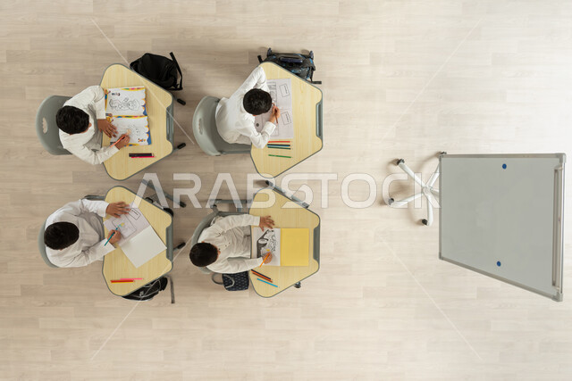 قرطاسية و أدوات مدرسية، انشطة ترفيهية ممتعة، بورتريه علوي عامودي لتلاميذ عرب خليجيون سعوديون يرتدون الثوب السعودي التقليدي يجلسون على الكراسي وأمامهم طاولات الدراسة، الترفيه وقضاء اوقات مسلية من خلال الرسم والتلوين، خلفية بيج