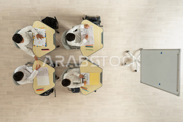 تنمية المواهب الفنية المسلية، بورتريه علوي عامودي لتلاميذ عرب خليجيون سعوديون يرتدون الثوب السعودي التقليدي يجلسون على الكراسي وأمامهم طاولات الدراسة، قضاء اوقات مسلية بالرسم والتلوين، خلفية بيج