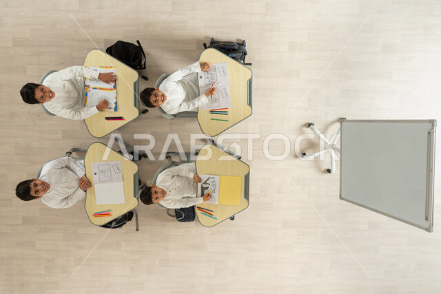 النظر الى الكاميرا بإيماءات تدل على السعادة والانجاز، بورتريه علوي عامودي لتلاميذ عرب خليجيون سعوديون يرتدون الثوب السعودي التقليدي يجلسون على الكراسي وأمامهم طاولات الدراسة، المساعدة والتعاون بين بعضهم البعض، قضاء اوقات مسلية بالرسم والتلوين،خلفية بيج