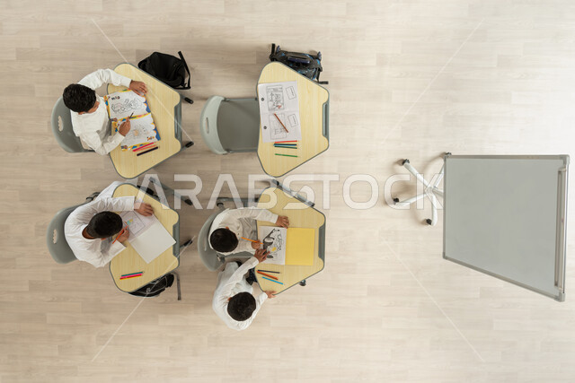 انشطة ترفيهية ممتعة ومسلية، قضاء اوقات مسلية بالرسم والتلوين، بورتريه علوي عامودي لتلاميذ عرب خليجيون سعوديون يرتدون الثوب السعودي التقليدي يجلسون على الكراسي وأمامهم طاولات الدراسة، المساعدة والتعاون بين بعضهم البعض، خلفية بيج