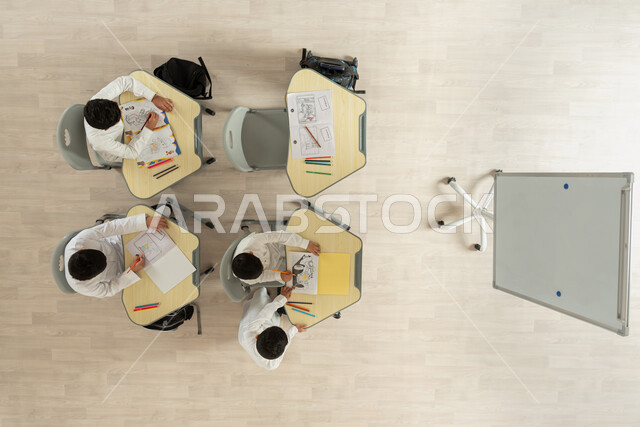 المساعدة والتعاون بين بعضهم البعض، قضاء اوقات مسلية بالرسم والتلوين، بورتريه علوي عامودي لتلاميذ عرب خليجيون سعوديون يرتدون الثوب السعودي التقليدي يجلسون على الكراسي وأمامهم طاولات الدراسة، خلفية بيج