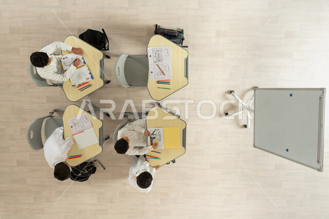 قضاء اوقات مسلية بالرسم والتلوين، بورتريه علوي عامودي لتلاميذ عرب خليجيون سعوديون يرتدون الثوب السعودي التقليدي يجلسون على الكراسي وأمامهم طاولات الدراسة، المساعدة والتعاون بين بعضهم البعض، خلفية بيج