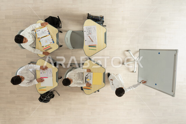 تنمية مهارة الرسم والتلوين، بورتريه علوي عامودي تلاميذ عرب خليجيون سعوديون يرتدون الثوب السعودي التقليدي يجلسون على الكراسي وأمامهم طاولات الدراسة، الانتباه لزميلهم الذي يرسم شيء ما على البورد الأبيض، خلفية بيج