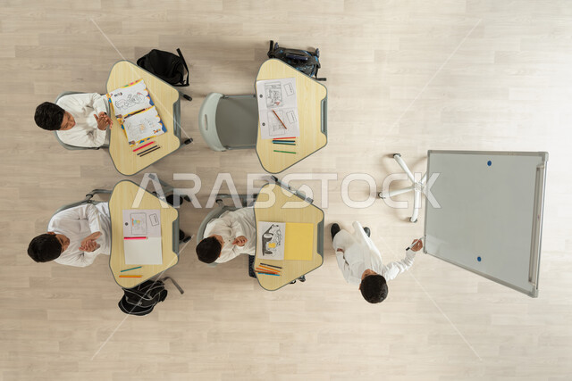 حب التعلم والتعليم، بورتريه علوي عامودي تلاميذ عرب خليجيون سعوديون يرتدون الثوب السعودي التقليدي يجلسون على الكراسي وأمامهم طاولات الدراسة، التصفيق لزميلهم الذي يرسم شيء ما على البورد الأبيض، تنمية مهارة الرسم والتلوين، خلفية بيج