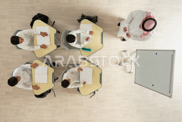 Following up on lessons and assignments, a vertical portrait of a Saudi Gulf Arab teacher giving lessons, Saudi Gulf Arab students wearing the traditional Saudi dress sitting on chairs with study tables in front of them, interest in science and learning, beige background
