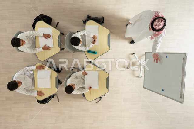 اكتساب المعرفة و الخبرات العلمية، بورتريه علوي عامودي لمعلم عربي خليجي سعودي يقوم بإعطاء الدروس، تلاميذ عرب خليجيون سعوديون يرتدون الثوب السعودي التقليدي يجلسون على الكراسي وأمامهم طاولات الدراسة، الانتباه الى الأستاذ لأنه يقوم برفع يده لشرح فكرة ما، خلفية بيج