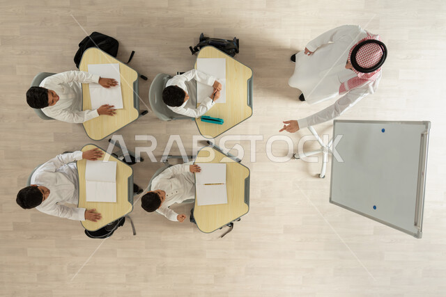 المثابرة على الدراسة لتحقيق النجاح، بورتريه علوي عامودي لمعلم عربي خليجي سعودي يقوم بإعطاء الدروس، تلاميذ عرب خليجيون سعوديون يرتدون الثوب السعودي التقليدي، الأستاذ يقوم برفع السبابة نحو الطالب ليجيب على السؤال المطروح، خلفية بيج