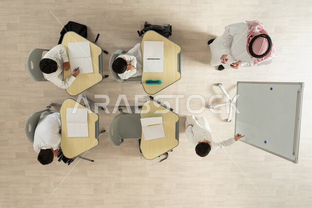 دورات تعليمية ثقافية، بورتريه علوي عامودي لمعلم عربي خليجي سعودي يقوم بإعطاء الدروس، تلاميذ عرب خليجيون سعوديون يرتدون الثوب السعودي التقليدي  يجلسون على الكراسي وأمامهم طاولات الدراسة، الانتباه للطالب الذي يشرح شيء ما من البورد الأبيض، خلفية بيج