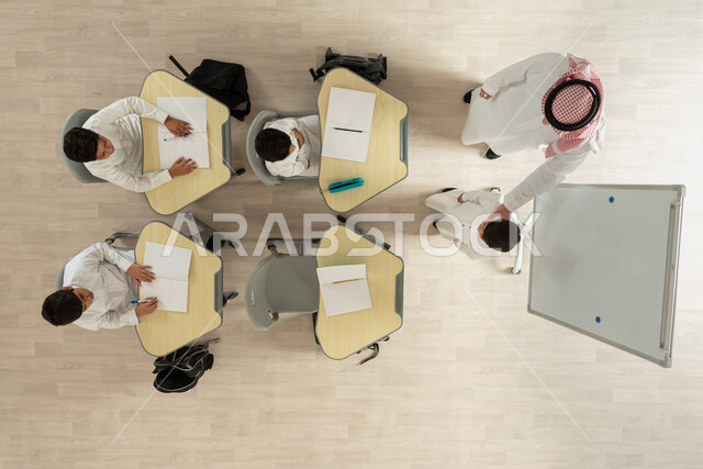 انشطة لتعليم الطلاب في المدارس، تلاميذ عرب خليجيون سعوديون يرتدون الثوب السعودي التقليدي  يجلسون على الكراسي وأمامهم طاولات الدراسة، بورتريه علوي عامودي لمعلم عربي خليجي سعودي يقوم بإعطاء الدروس، الاهتمام بالعملية التعليمية، خلفية بيج