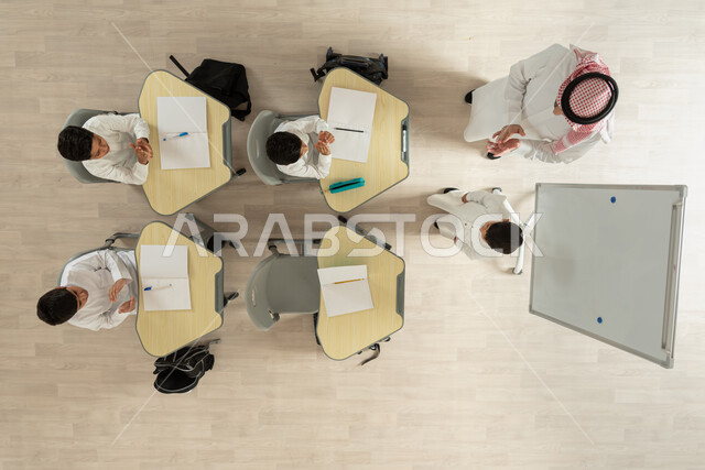 الانجاز و التفوق في المرحلة التعليمية، تلاميذ عرب خليجيون سعوديون يرتدون الثوب السعودي التقليدي  يجلسون على الكراسي وأمامهم طاولات الدراسة، بورتريه علوي عامودي لمعلم عربي خليجي سعودي يقوم بإعطاء الدروس، التصفيق للطالب المتميز، خلفية بيج