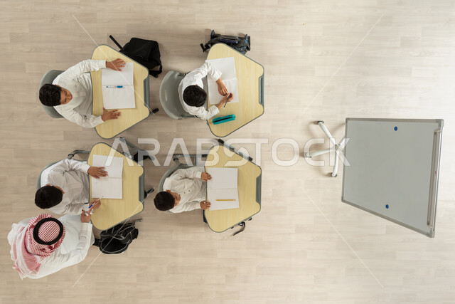 حل الواجبات اليومية، تلاميذ عرب خليجيون سعوديون يرتدون الثوب السعودي التقليدي  يجلسون على الكراسي وأمامهم طاولات الدراسة، بورتريه علوي عامودي لمعلم عربي خليجي سعودي يقوم بمساعدة الطلاب، الاهتمام بالتعلم و التعليم، خلفية بيج