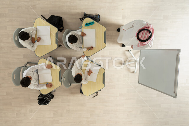 الاهتمام بتعليم الطلاب و تنمية القدرات العلمية، تأدية الواجبات المدرسية، تلاميذ عرب خليجيون سعوديون يرتدون الثوب السعودي التقليدي  يجلسون على الكراسي وأمامهم طاولات الدراسة، بورتريه علوي عامودي لمعلم عربي خليجي سعودي يقوم بإعطاء الدروس، خلفية بيج