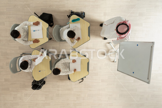 تأدية الواجبات المدرسية، تنمية القدرات الإبداعية لدى الطلاب، تلاميذ عرب خليجيون سعوديون يرتدون الثوب السعودي التقليدي  يجلسون على الكراسي وأمامهم طاولات الدراسة، بورتريه علوي عامودي لمعلم عربي خليجي سعودي يقوم بإعطاء الدروس، الاهتمام بالعلم والتعلم، خلفية بيج