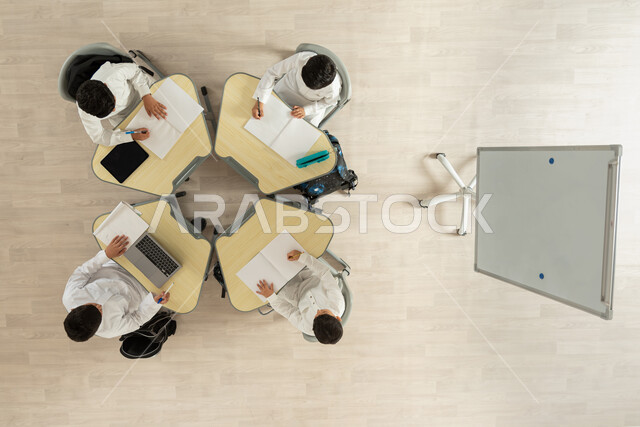 تنمية القدرات الإبداعية لدى الطلاب، تأدية الواجبات المدرسية، بورتريه علوي عامودي لتلاميذ عرب خليجيون سعوديون يرتدون الثوب السعودي التقليدي  يجلسون على الكراسي وأمامهم طاولات الدراسة، الاهتمام بالعلم والتعلم، دمج العملية التعليمية مع التكنولوجية، خلفية بيج